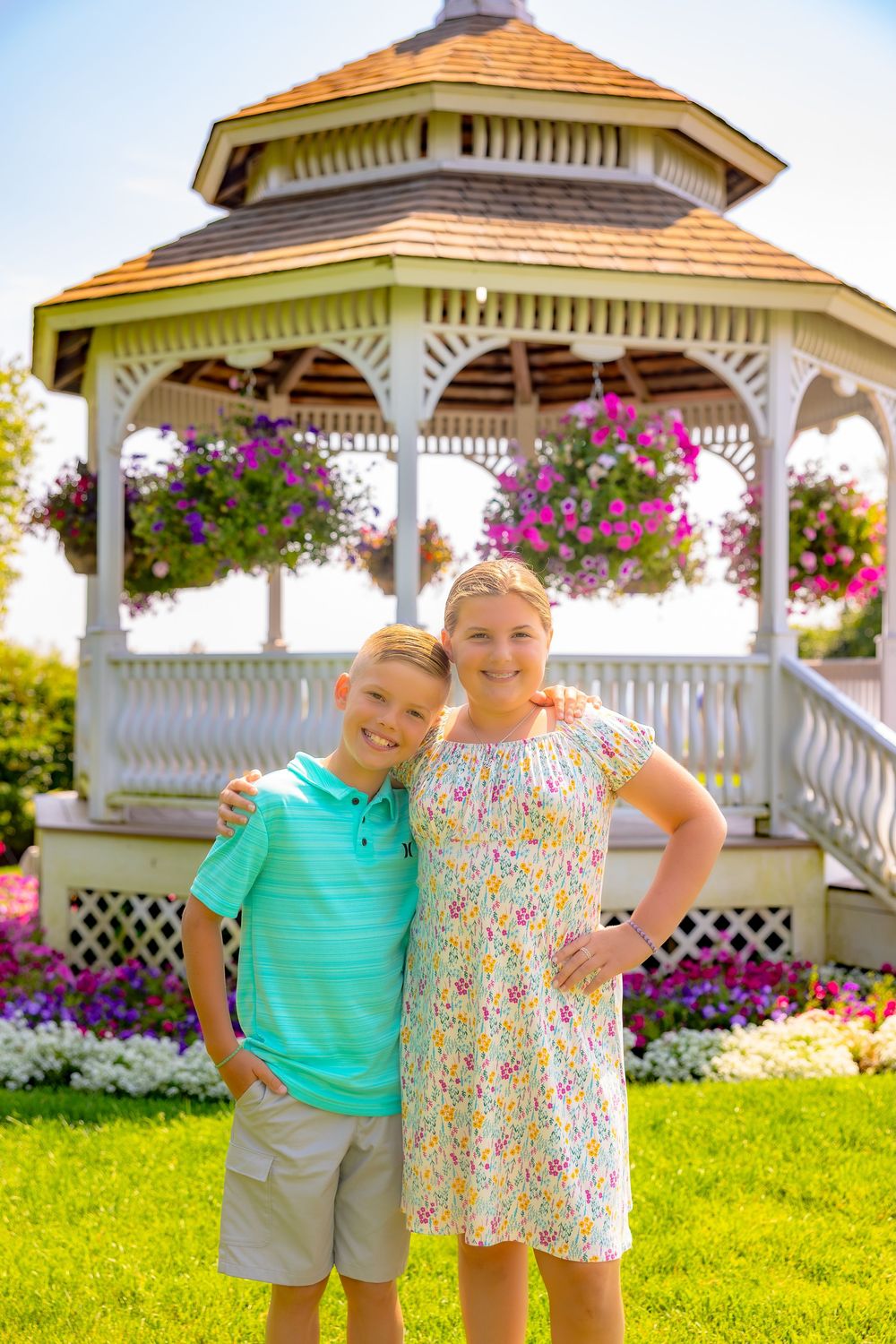 Two children stand together near a white gazebo surrounded by colorful hanging flower baskets and green grass.