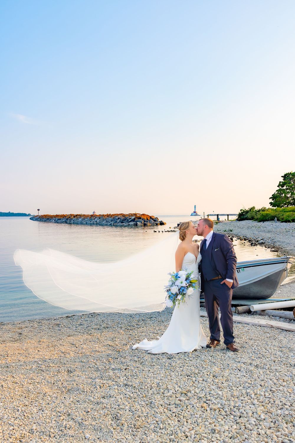 A romantic wedding couple shares a tender moment on a rocky beach at sunset with boats in the background.