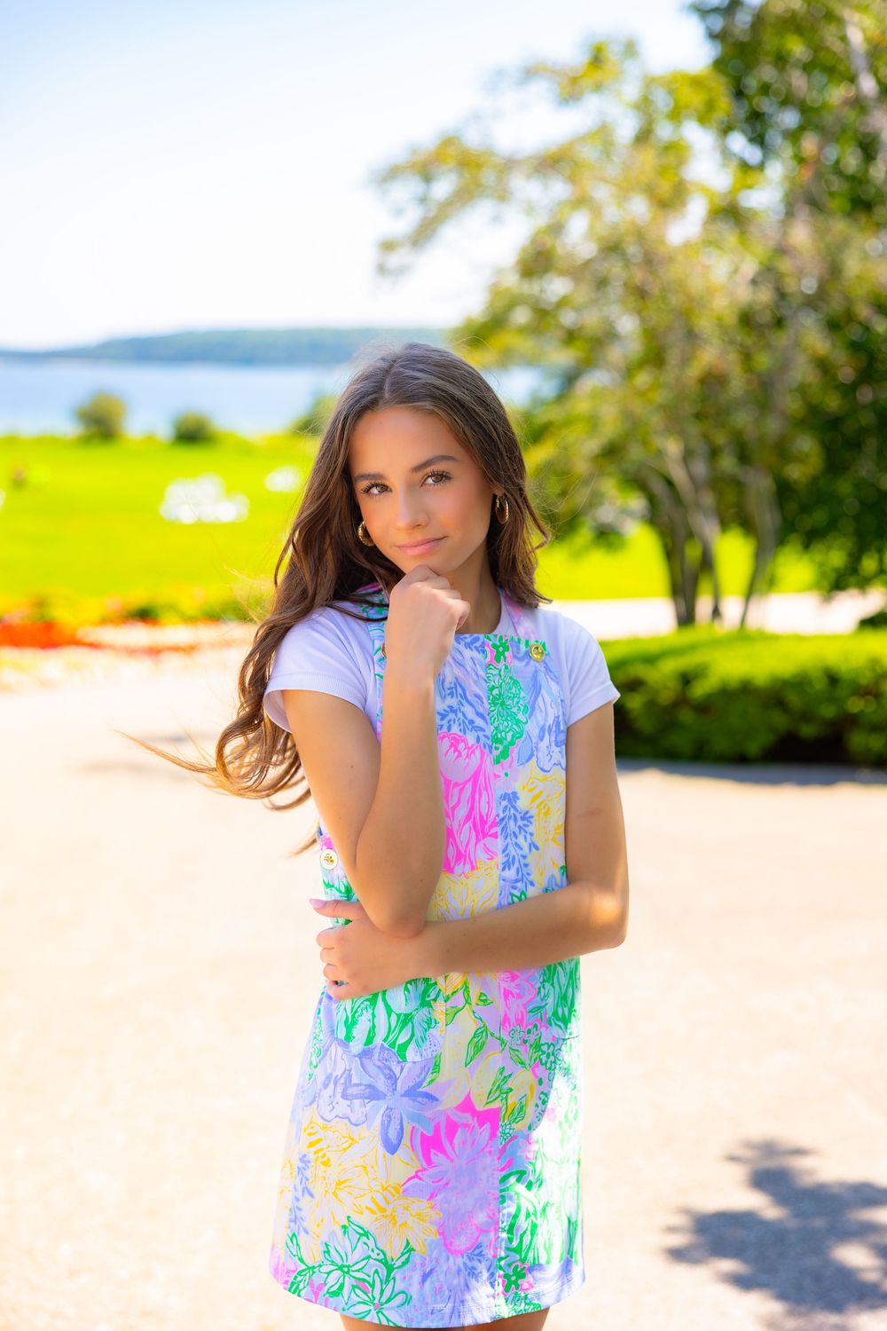 A young person in a colorful floral print t-shirt poses outdoors on a sunny day with trees and water in the background.