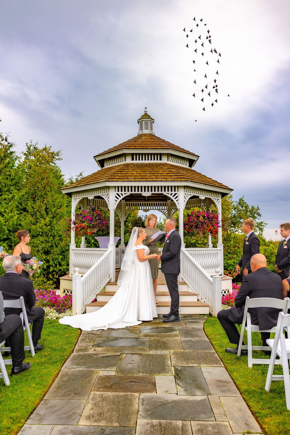 An outdoor wedding ceremony takes place under a white gazebo with guests seated on white chairs and cloudy skies above.