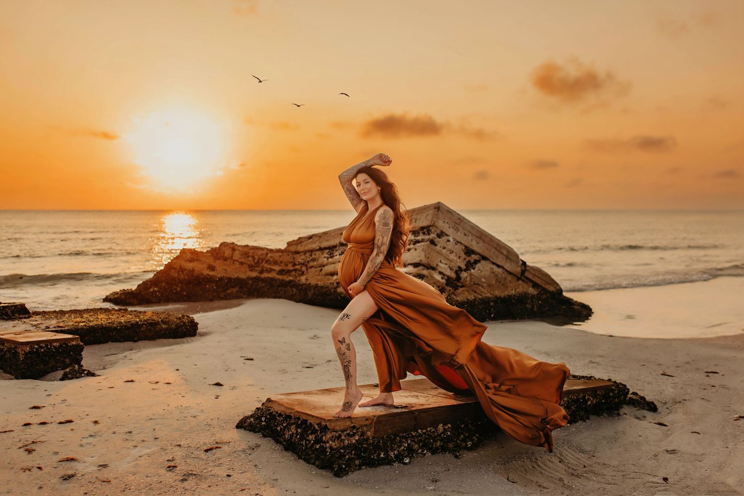 Woman in flowing rust-colored dress poses on beach rocks during golden hour sunset.