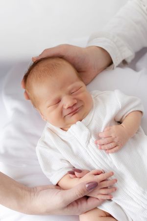 Smiling baby in white clothing lying on white bedding held by loving hands.