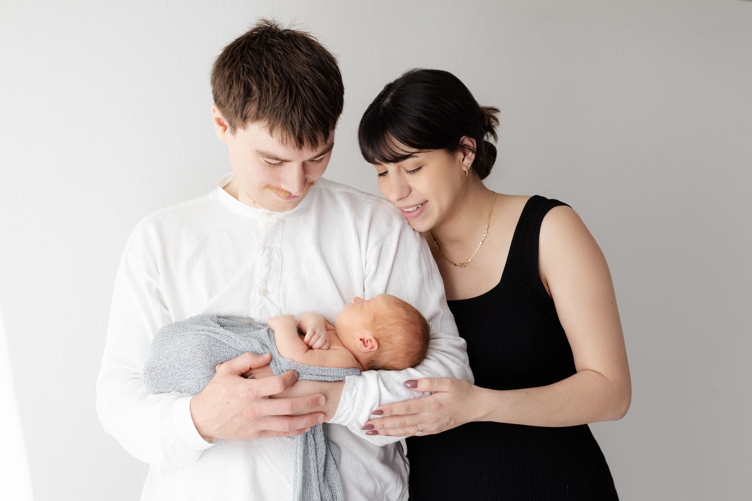 A couple in white and black clothing tenderly holding their newborn baby against a light colored background.