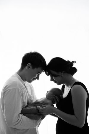 Silhouette portrait shows couple tenderly gazing down at their newborn baby against white background.