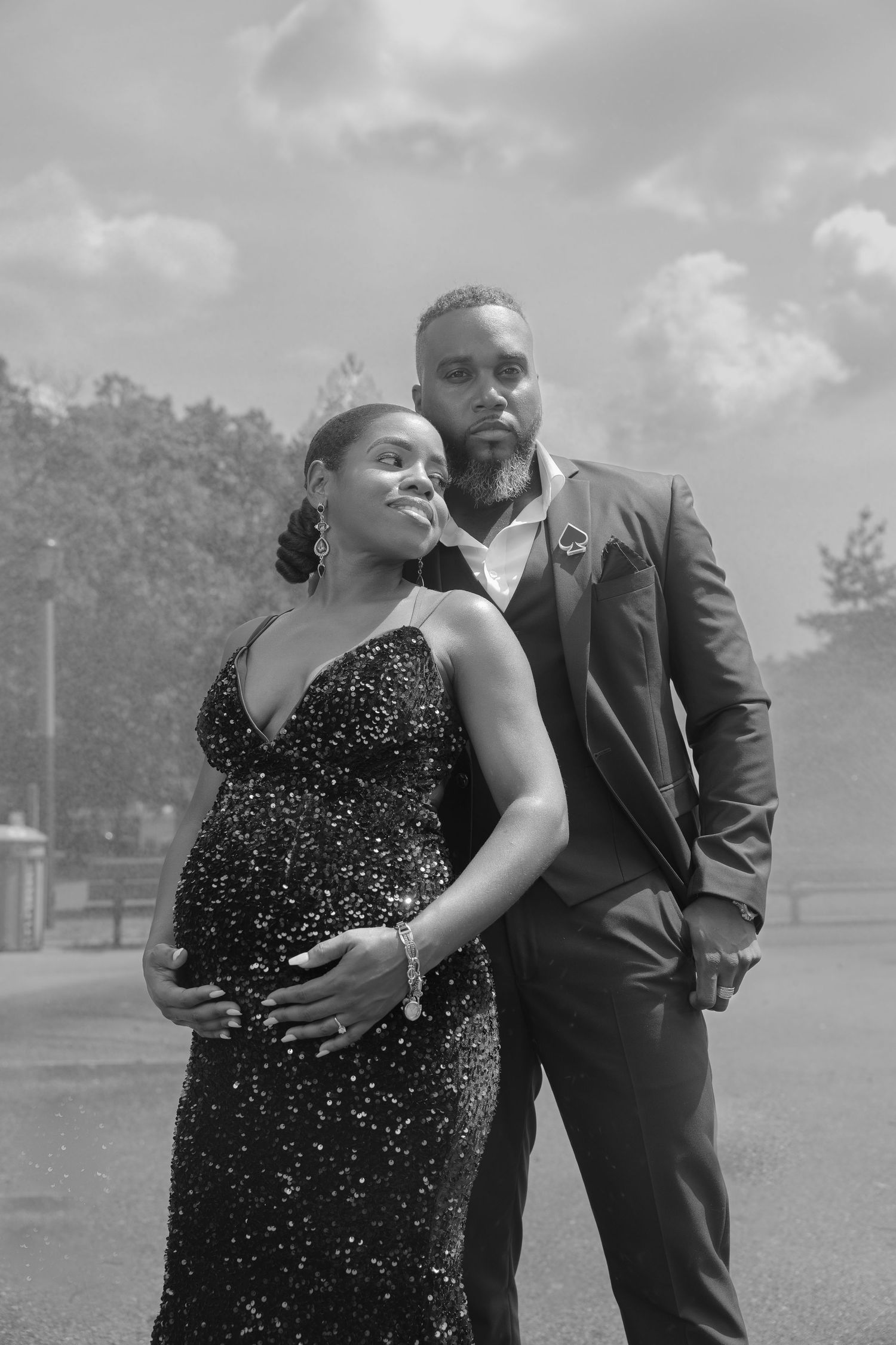 A black and white photo of a couple in formal attire embracing outdoors, with her in a sparkly dress showing a baby bump.