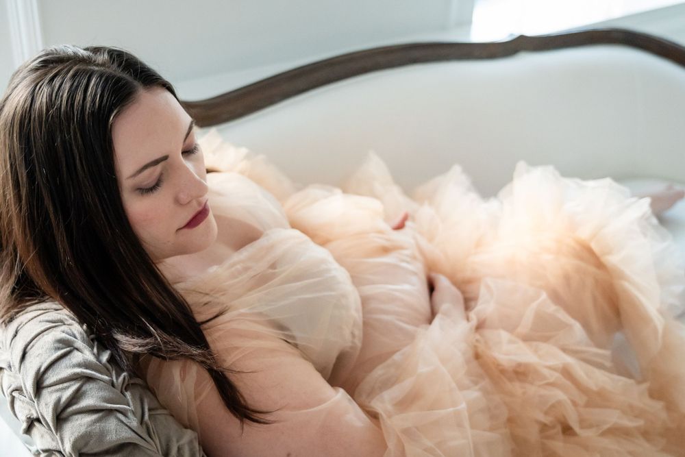 Ethereal maternity portrait with woman in flowing tulle dress resting on couch.