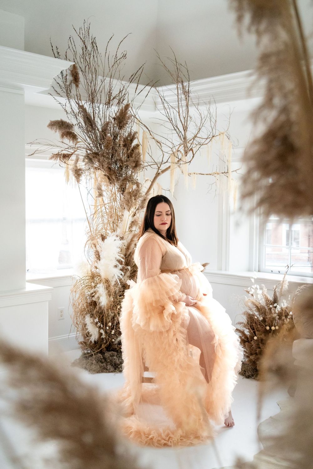 A maternity photo shoot featuring a flowing peach tulle gown and dried floral arrangements in a bright white room.