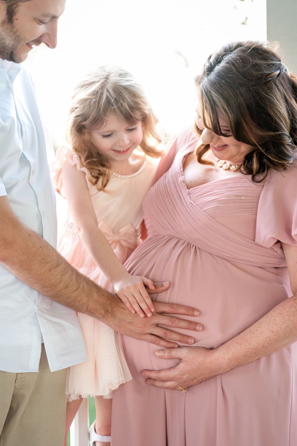 Tender family moment as they touch a pregnant belly in a pink maternity dress and white shirt.