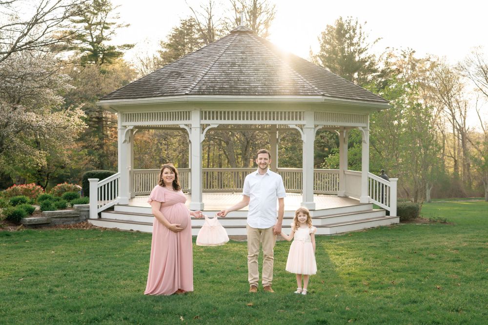Family in coordinated pink and white outfits pose together in front of a white gazebo at sunset.