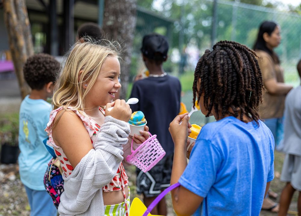 Two children smiling and enjoying ice cream treats outdoors at a summer camp event with other kids in the background.