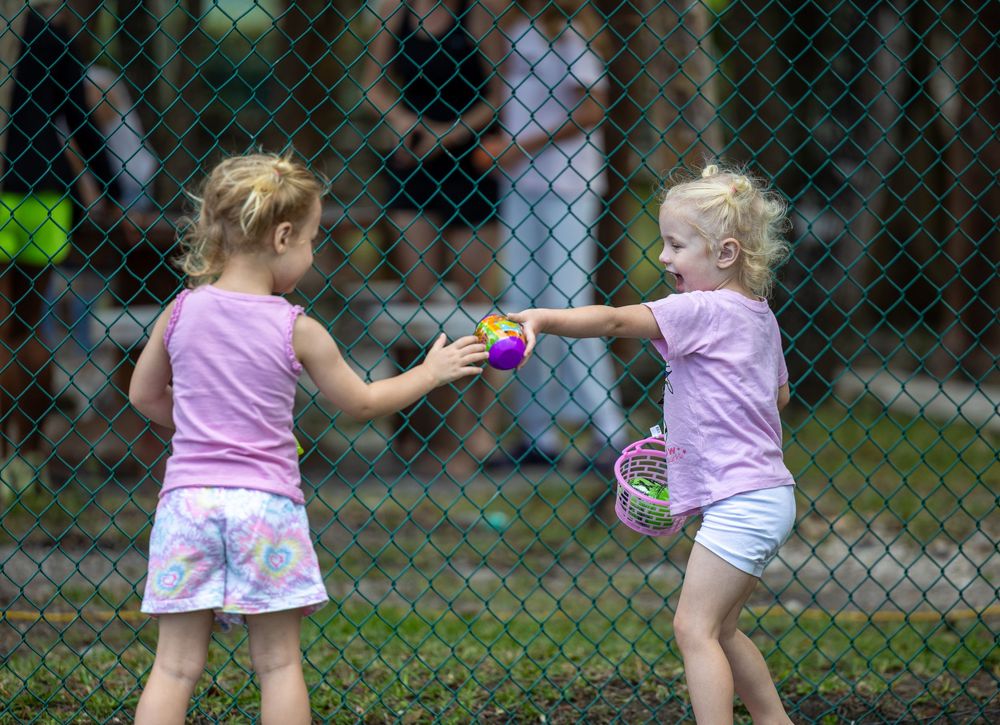 Two young girls in pink shirts exchange Easter eggs near a chain-link fence outdoors.