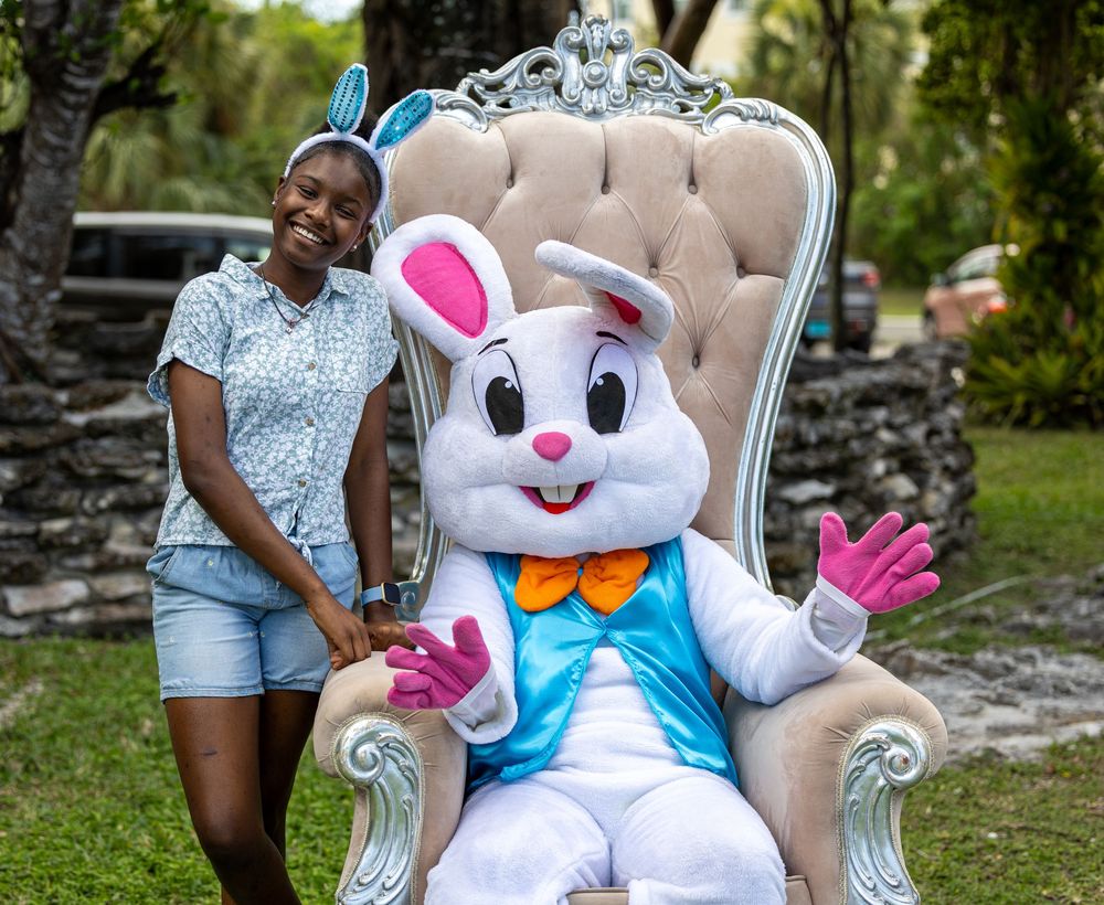 A smiling woman poses with the Easter Bunny mascot seated on an ornate white throne chair outdoors.
