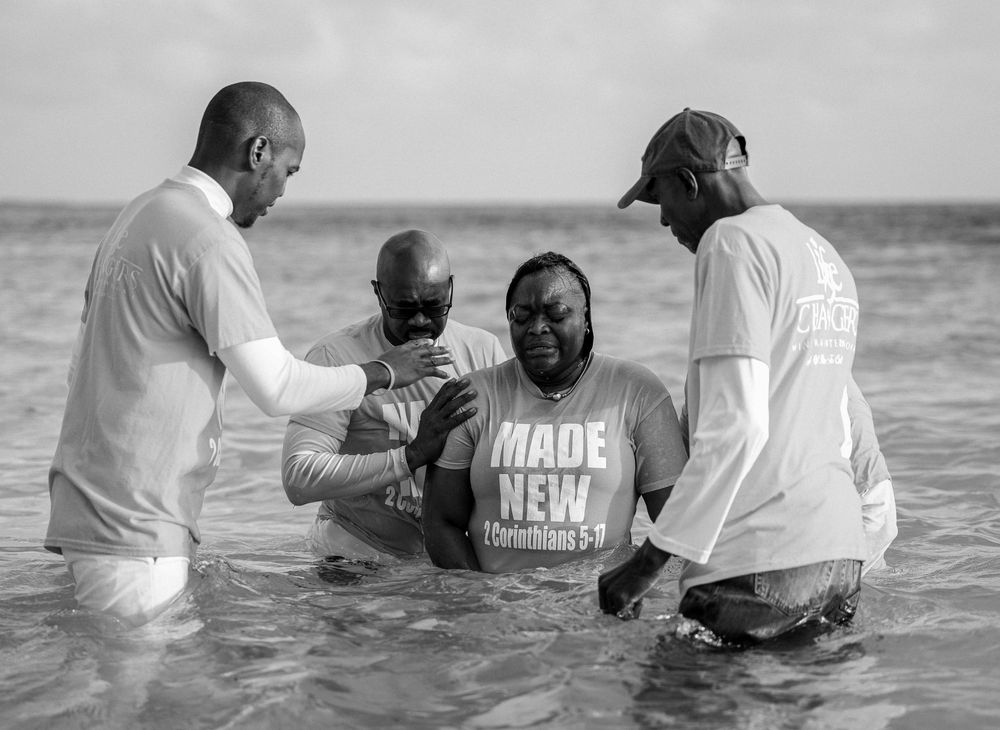 Group baptism ceremony in ocean waters, person wearing 'Made New' shirt surrounded by ministers.