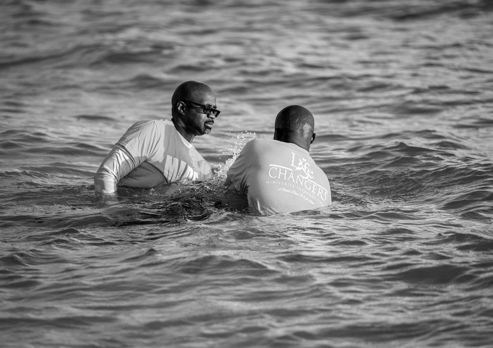 Two men in white shirts stand waist-deep in ocean water during a baptism ceremony in black and white photo.