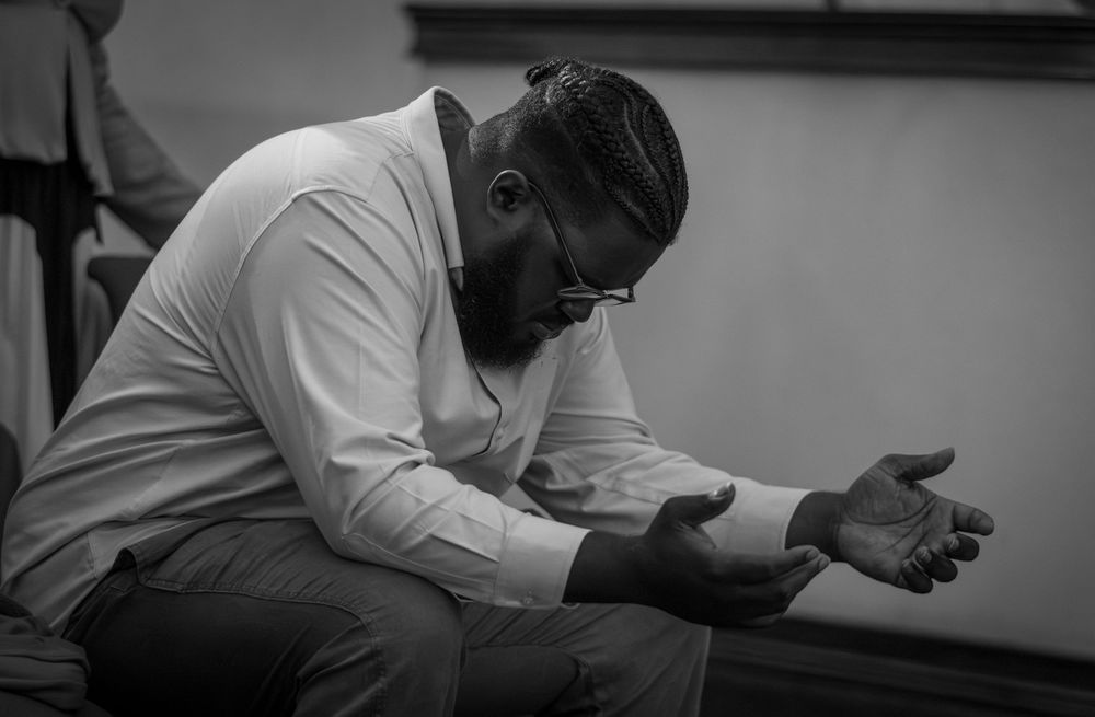 A person sits in deep contemplation with hands clasped in a black and white portrait.