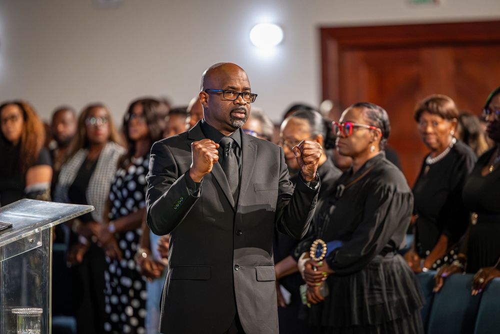 Man in black suit speaking passionately at a funeral service with mourners dressed in black standing behind him.