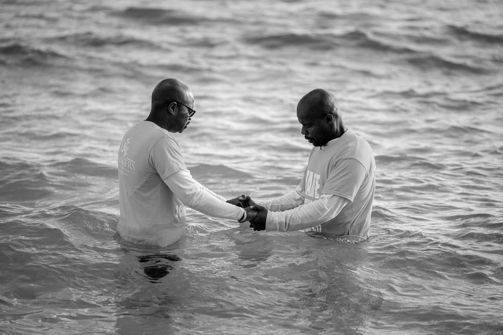 Two men in white shirts stand in ocean waves during a baptism ceremony, black and white photo.