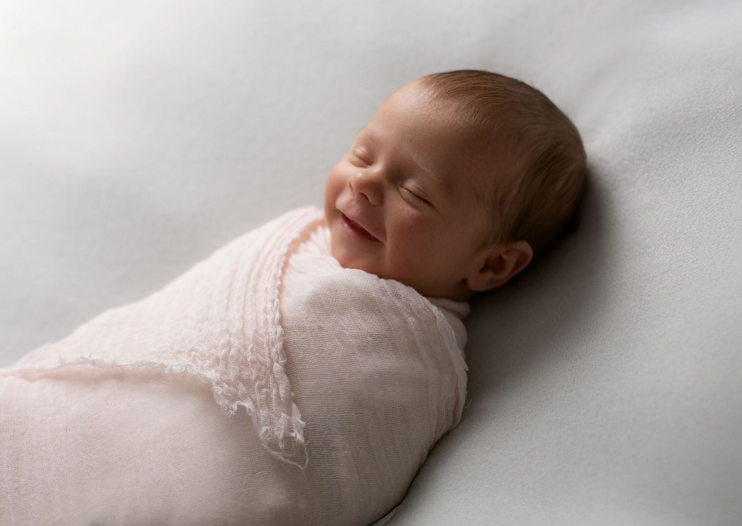 A newborn baby wrapped in soft white lace fabric smiles peacefully while sleeping.