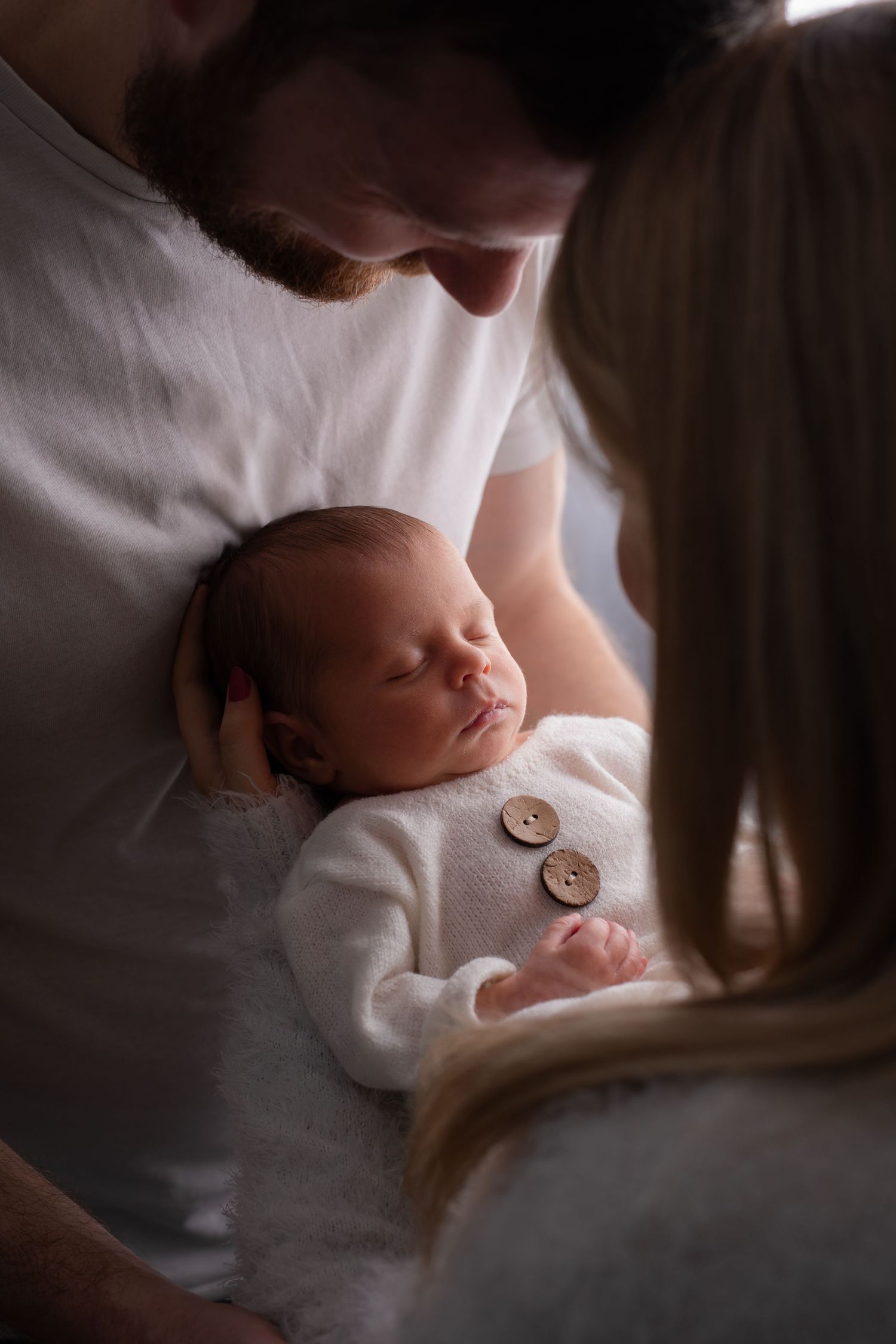 A sleeping newborn baby in white clothing being tenderly held while two adults gaze down lovingly.