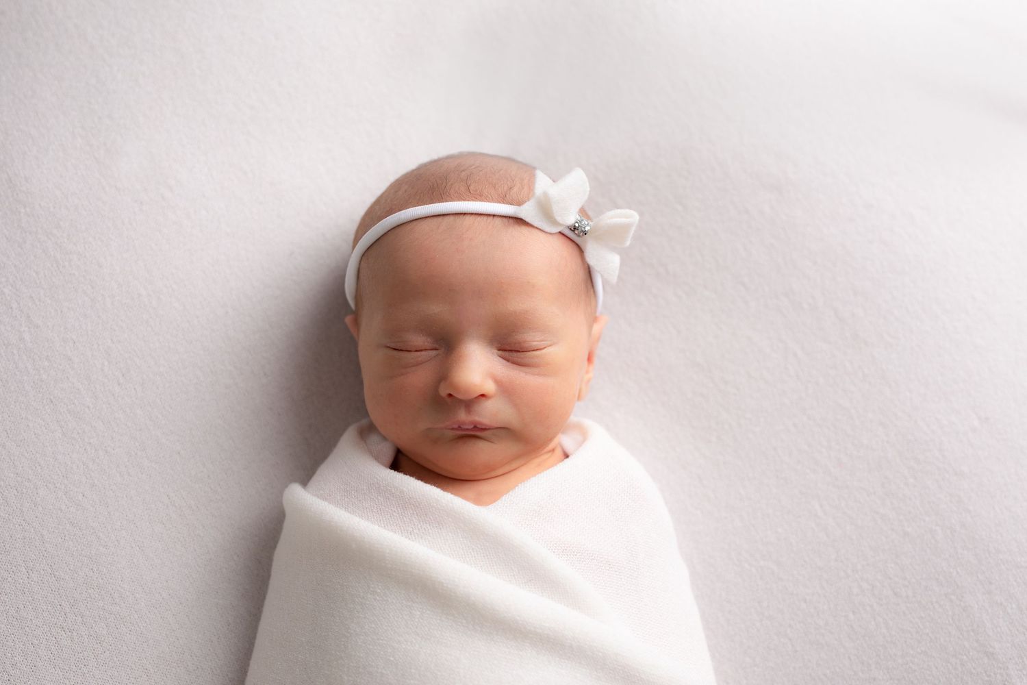 Newborn baby sleeps peacefully while wrapped in a white blanket, wearing a delicate floral headband.
