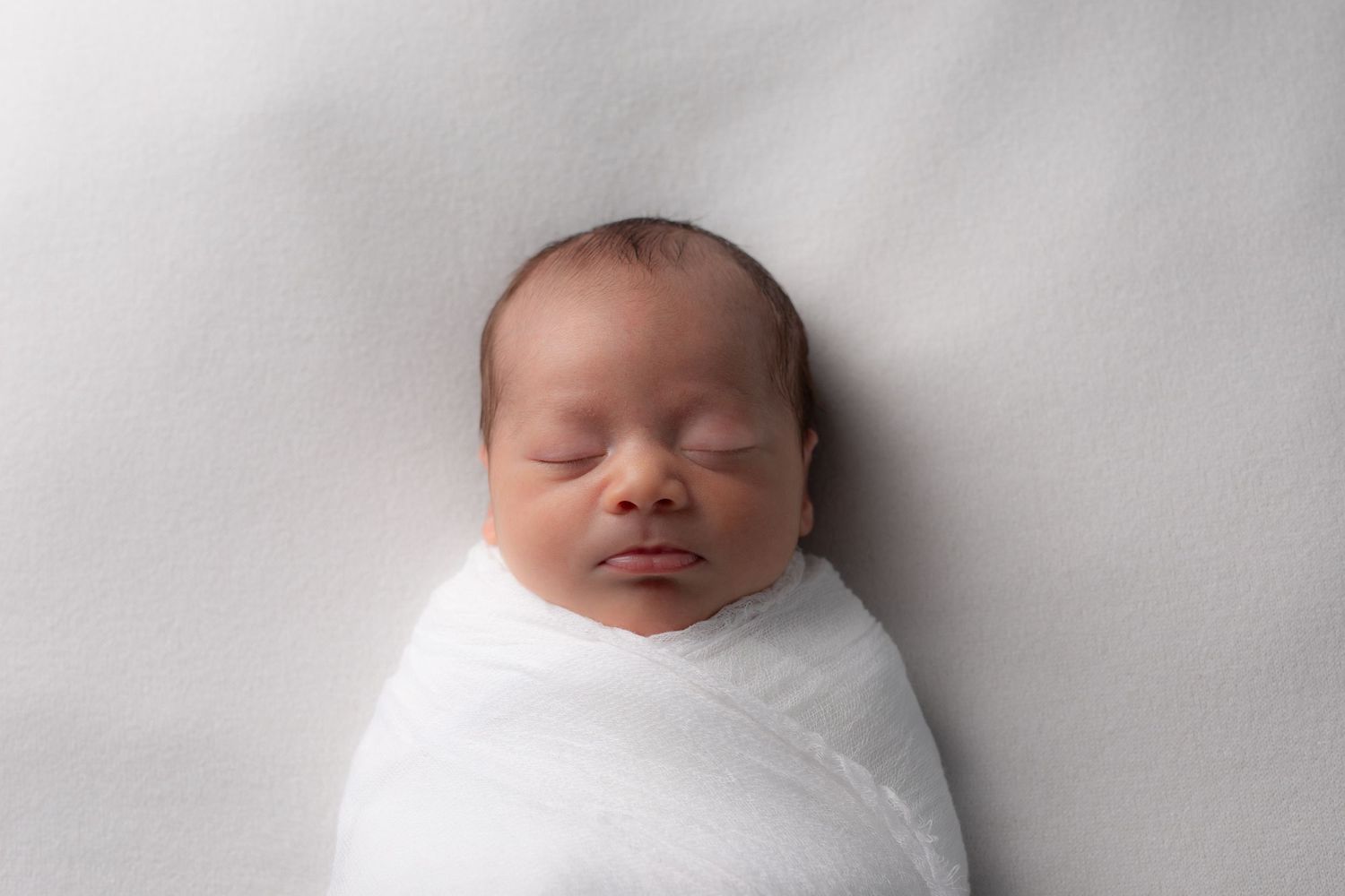 Peaceful sleeping newborn baby wrapped in soft white swaddle against light grey background.