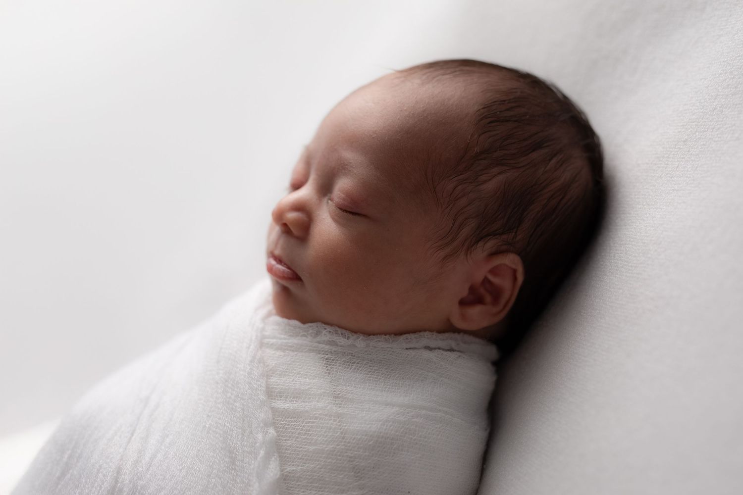 A peaceful sleeping newborn baby swaddled in soft white fabric against a white background.