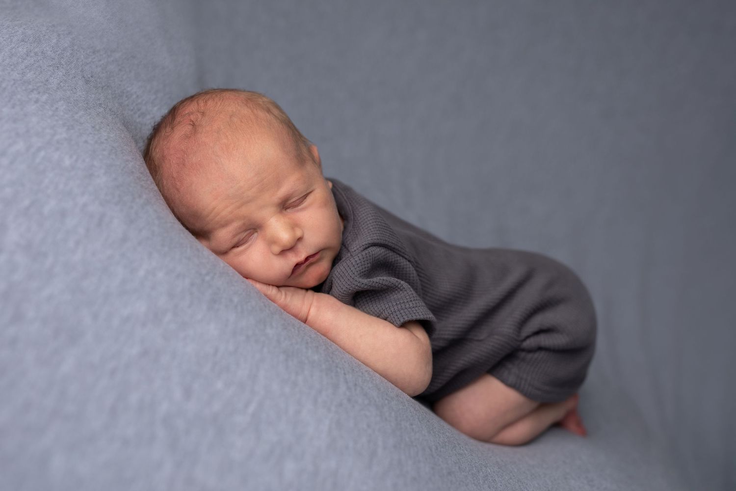Sleeping newborn baby wearing gray outfit curled up on soft gray backdrop in professional studio portrait series.