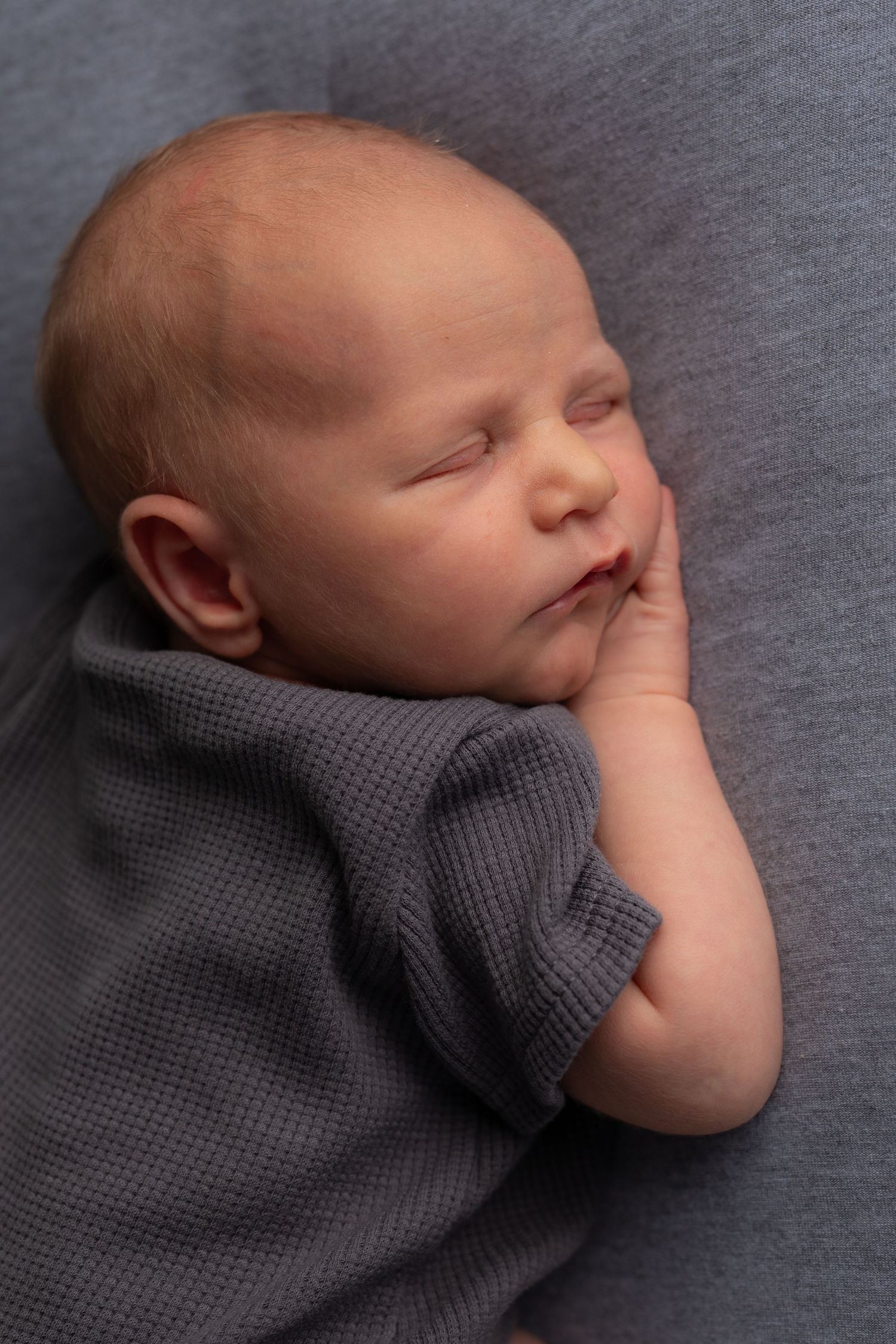 Peaceful sleeping newborn baby in gray knit clothing rests with hand near face on soft gray background.