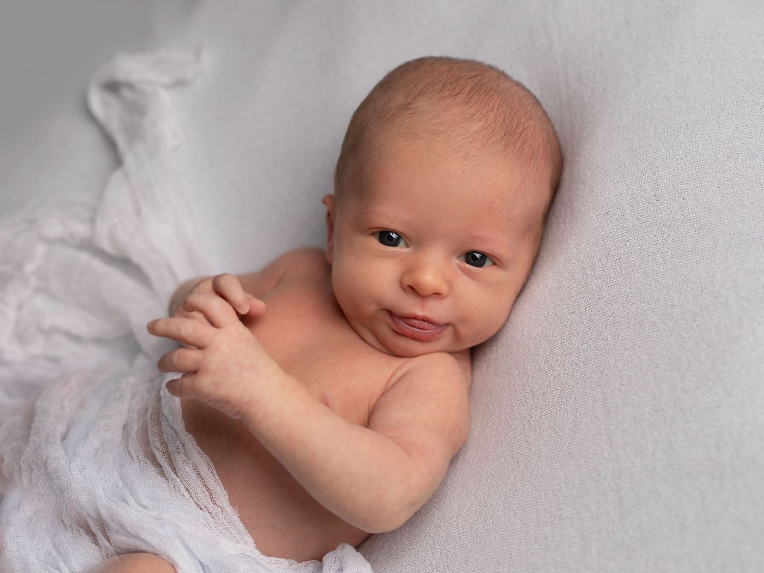 Newborn baby lying on white blanket with hand clasped and sweet expression.