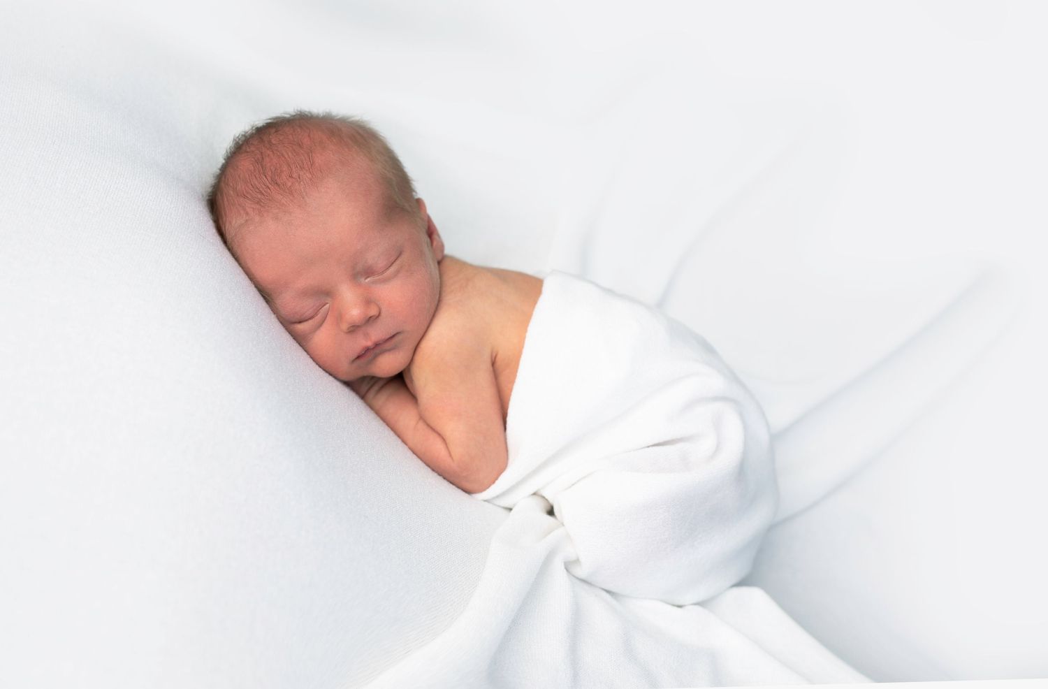 A sleeping newborn baby swaddled in white blankets rests peacefully on white bedding.
