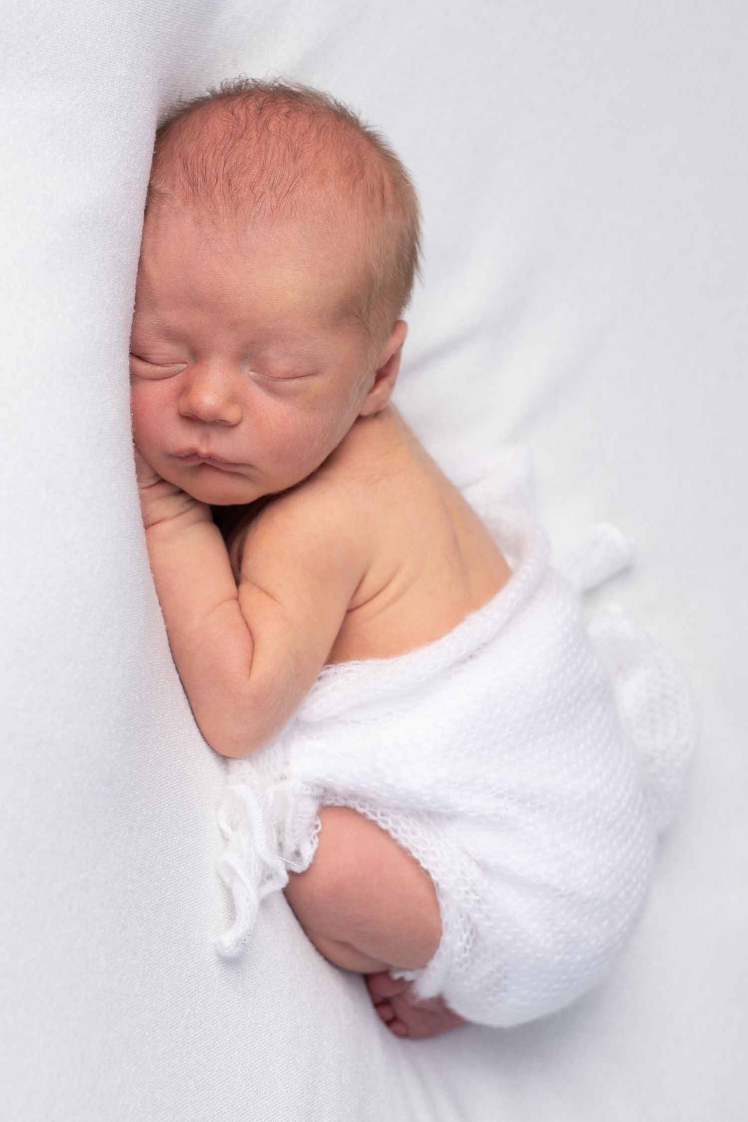 Sleeping newborn baby wrapped in white fabric against a light background.