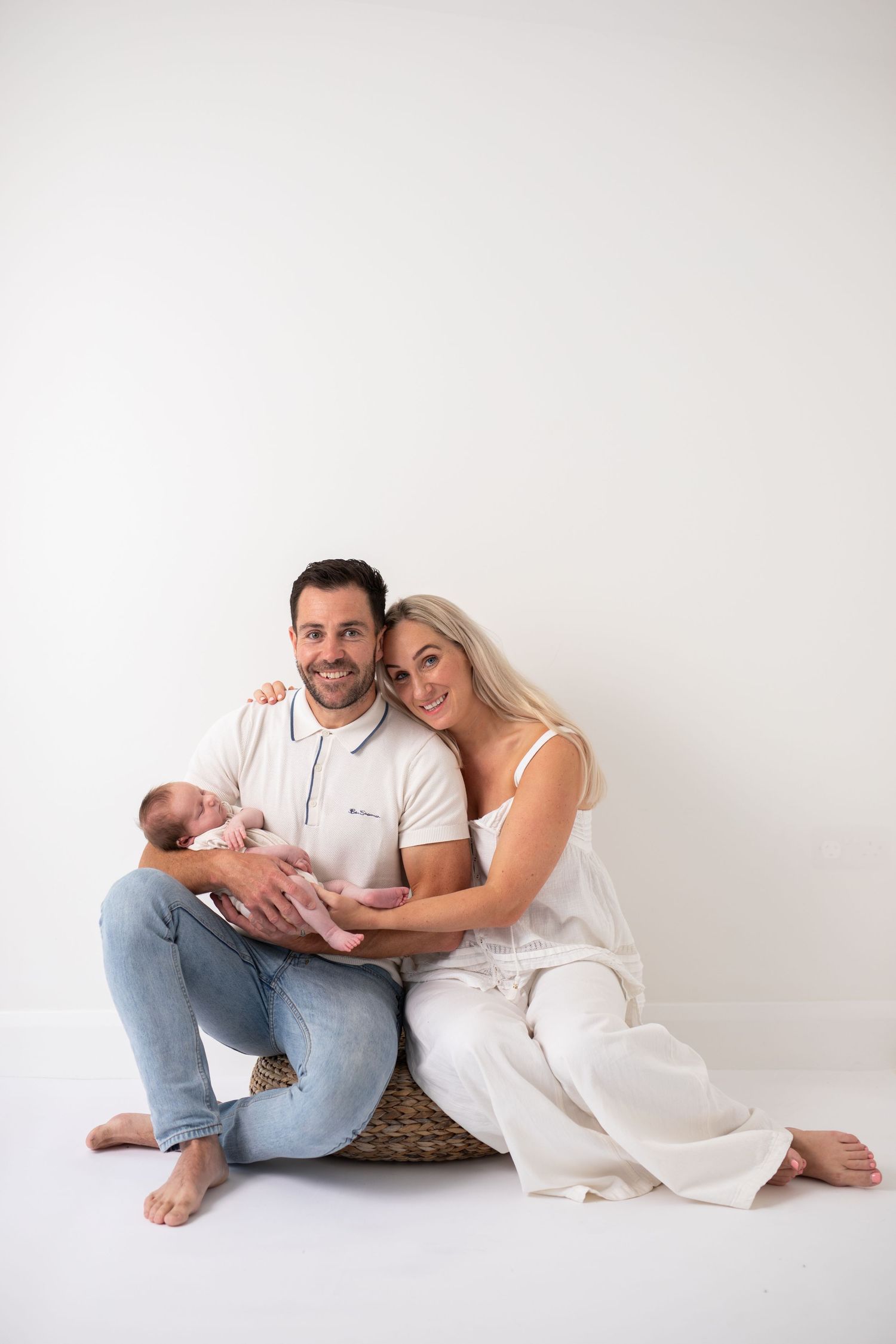 A couple in white and denim sit intimately together against a white backdrop in a casual studio portrait series.
