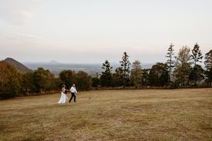 Couple in wedding attire poses on a scenic hilltop with mountains and trees in the background at golden hour.