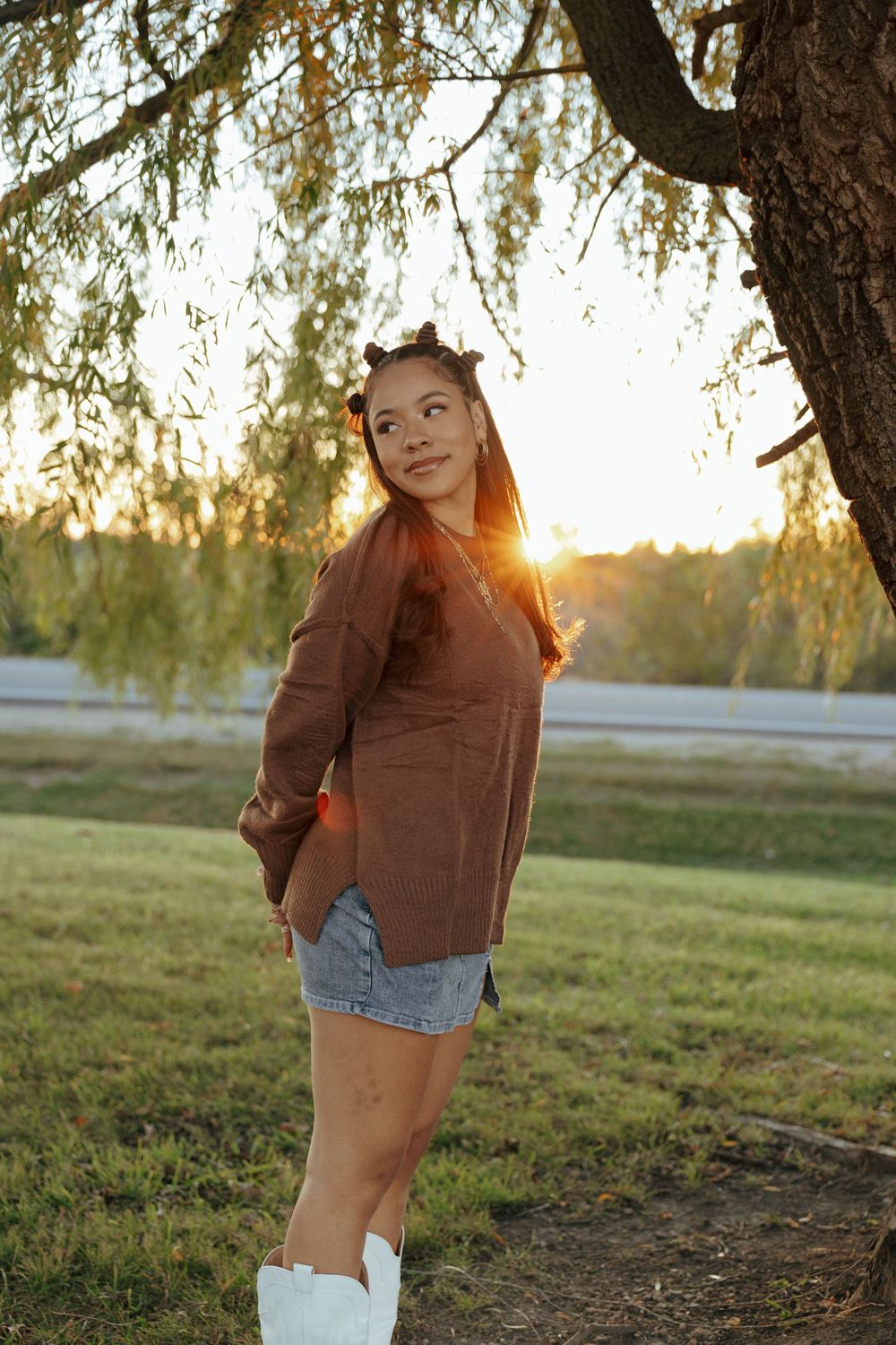 Person in denim shorts and brown sweater poses playfully in a grassy field at sunset.