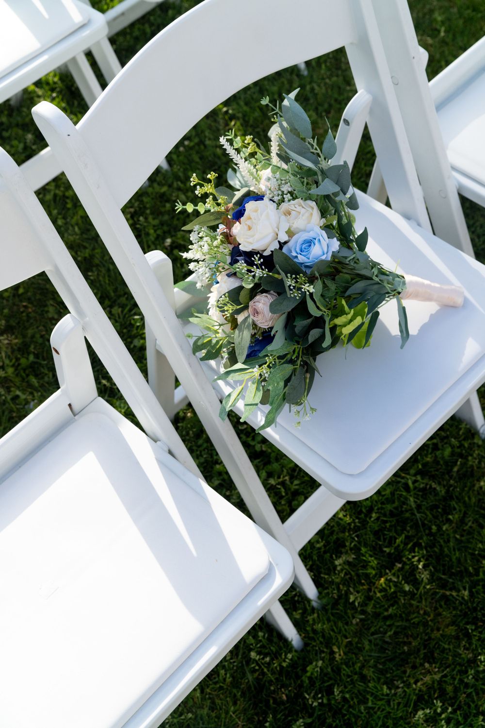 A delicate blue and white floral bouquet rests on a white folding chair on grass.