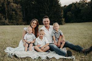 Family of five sits on white blanket in grassy field, parents with three young boys in white outfits.