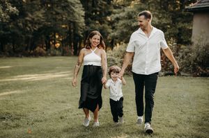 Young family of three walks hand-in-hand across green lawn, parents swinging toddler boy between them.