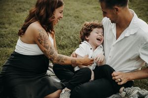Parents laughing with curly-haired toddler boy sitting between them on blanket outdoors in grass.