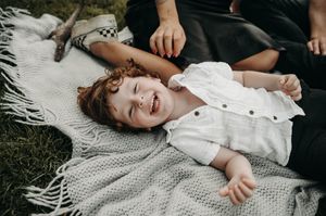 Laughing curly-haired toddler lies on cozy fringed blanket outdoors while parents sit nearby.