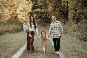 Happy family of four walks a wooded autumn trail, parents holding hands with two young children.