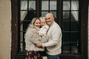 Smiling family of three posing together in front of vintage windows during an indoor photo session.