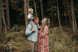 Family of three poses in forest, dad with toddler on shoulders, mom in floral dress smiling up at child.