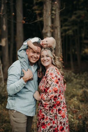 Happy family portrait in woods, dad holding child on back, mom in red floral dress embracing together.