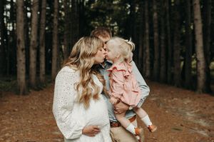 Mother and father share a kiss over smiling baby in pink shirt among pine trees on dirt path.