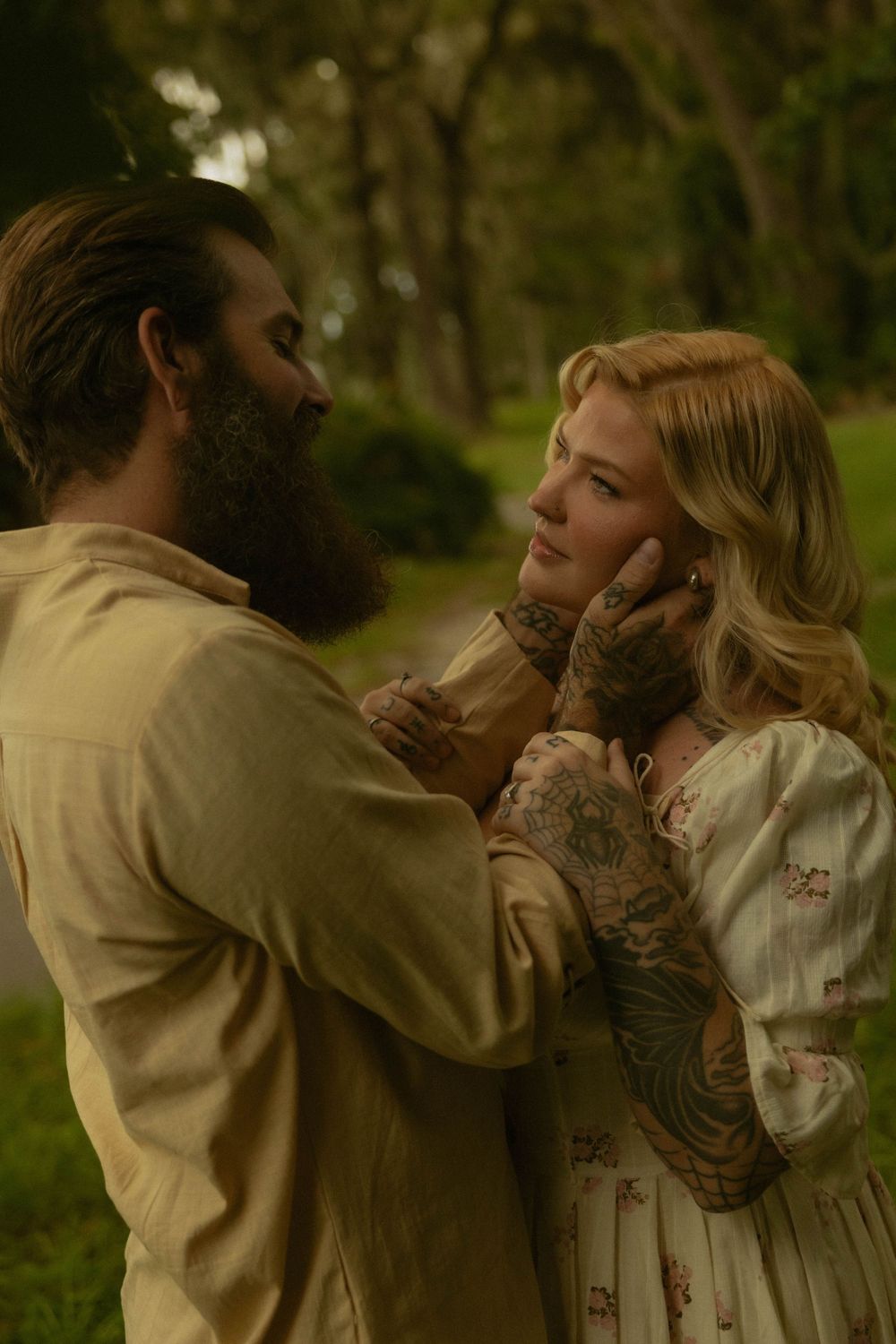 Tattooed bearded man tenderly holds blonde woman's face in a romantic outdoor garden setting.