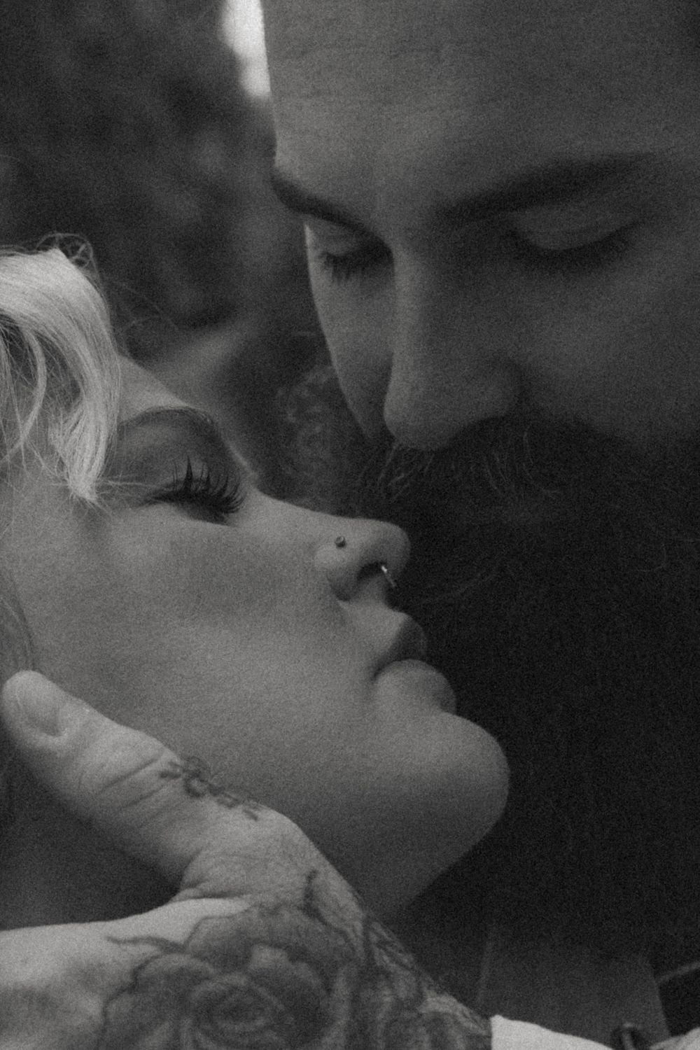 Black and white photo of a bearded man with tattoos leaning in close to a blonde woman with a nose ring.