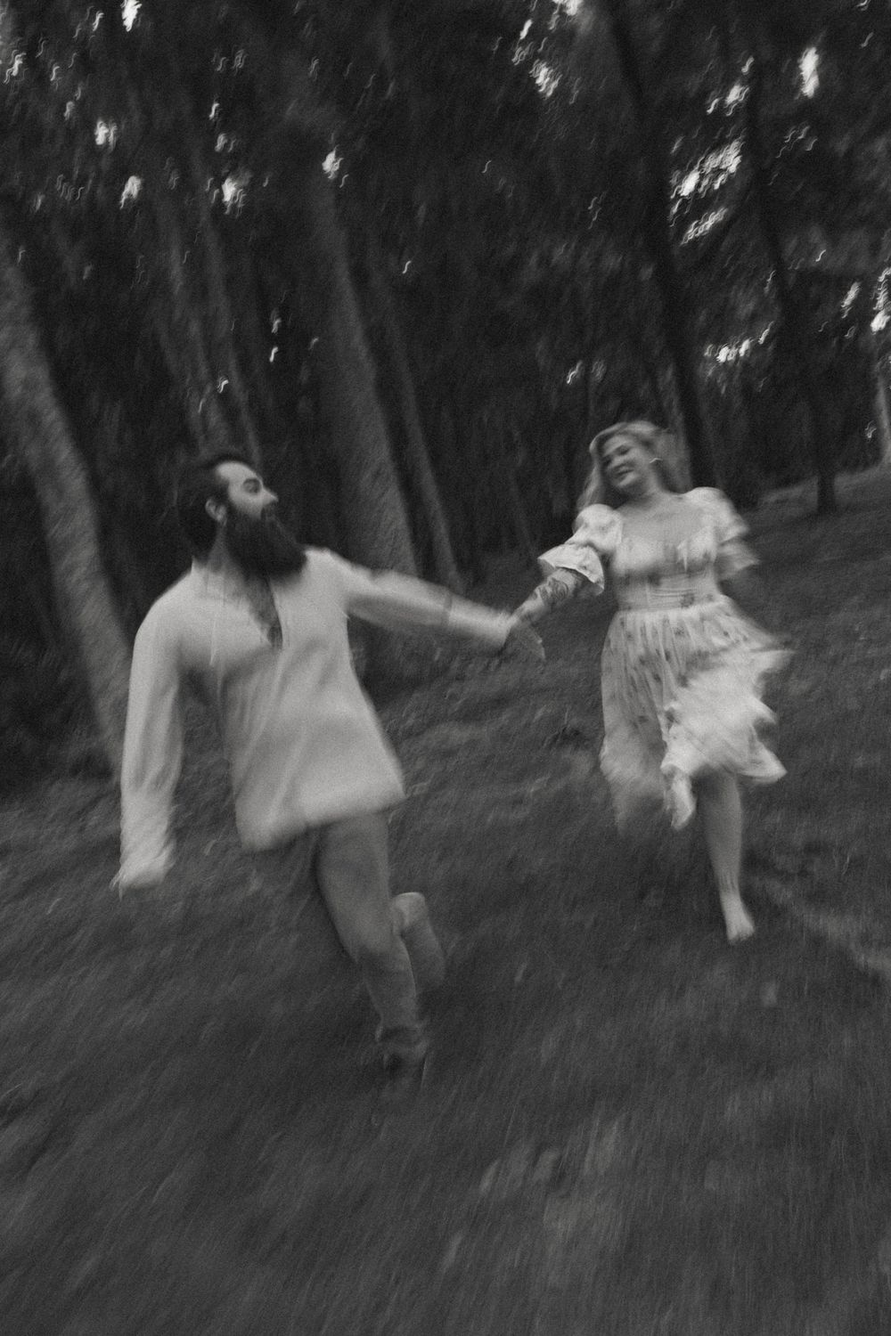 Black and white photo of a couple holding hands running through a forest, motion blur capturing their joyful movement.