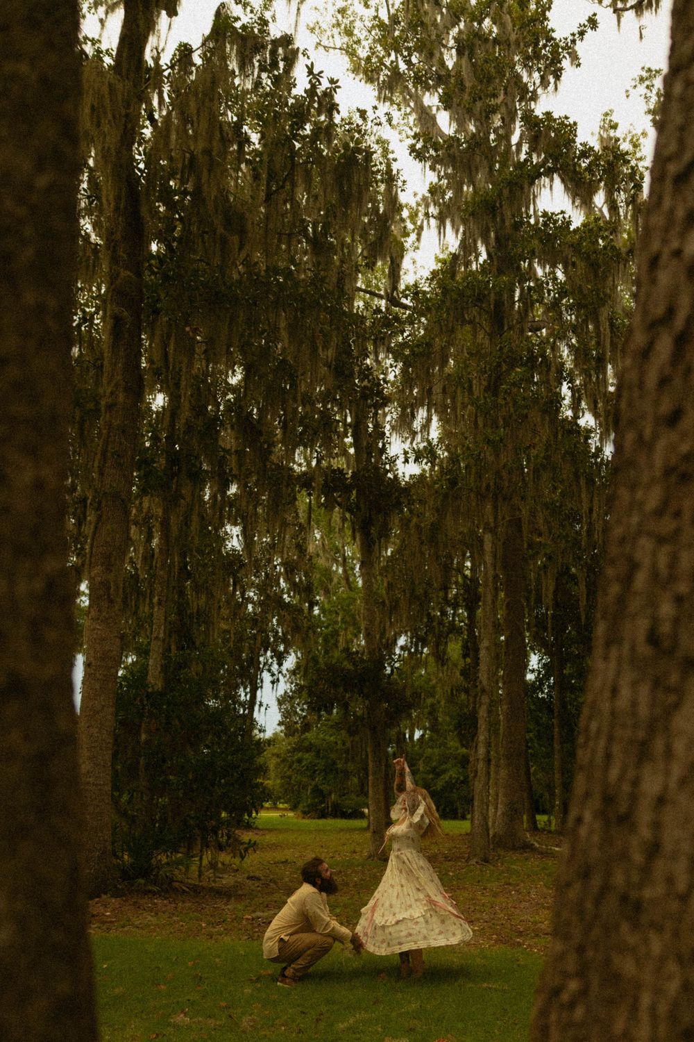 Couple dancing under Spanish moss-draped trees in a lush green park, woman in floral dress twirling gracefully.