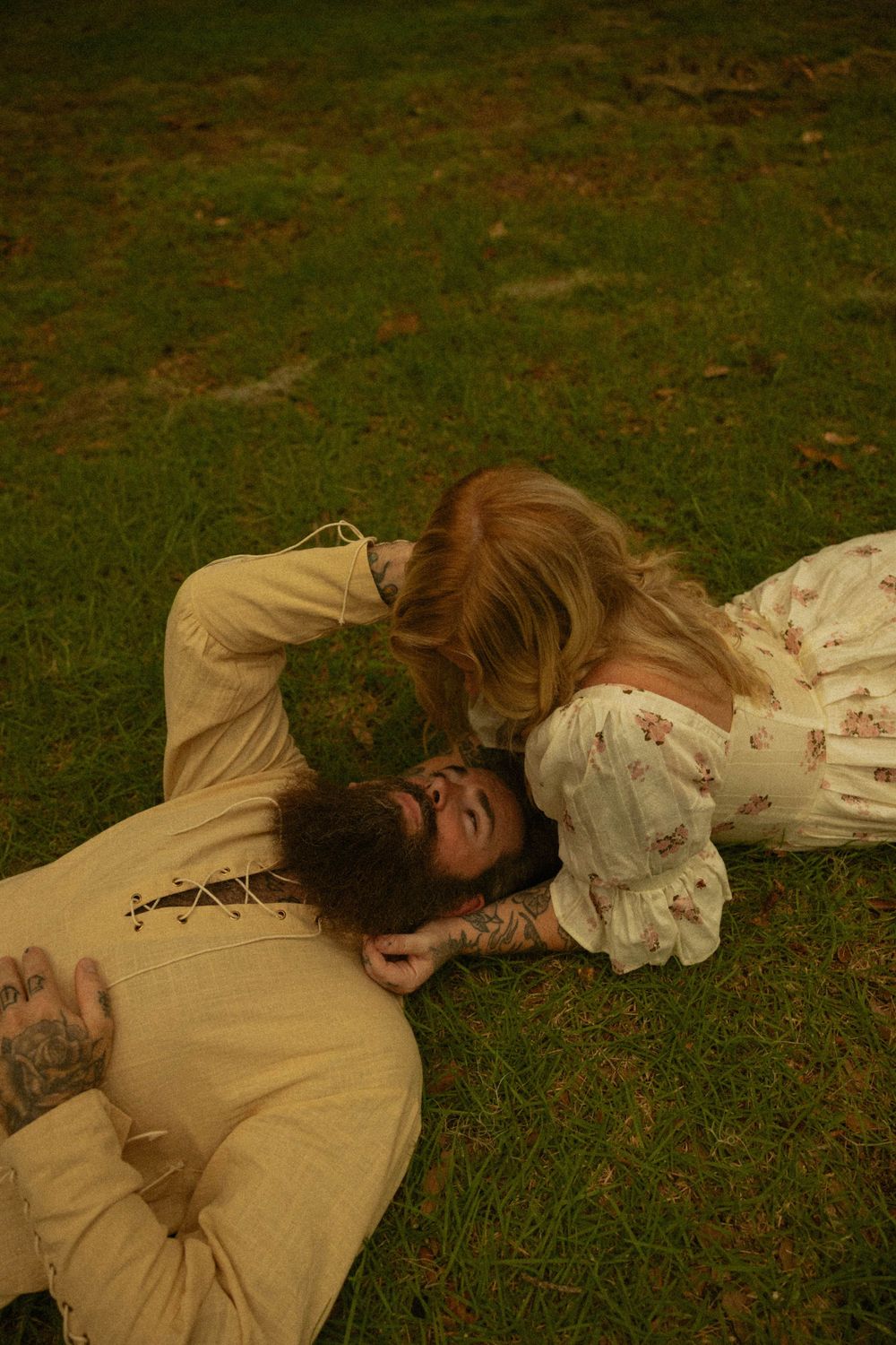 A bearded tattooed man and blonde woman in vintage floral dress lying on green grass in a romantic outdoor pose.