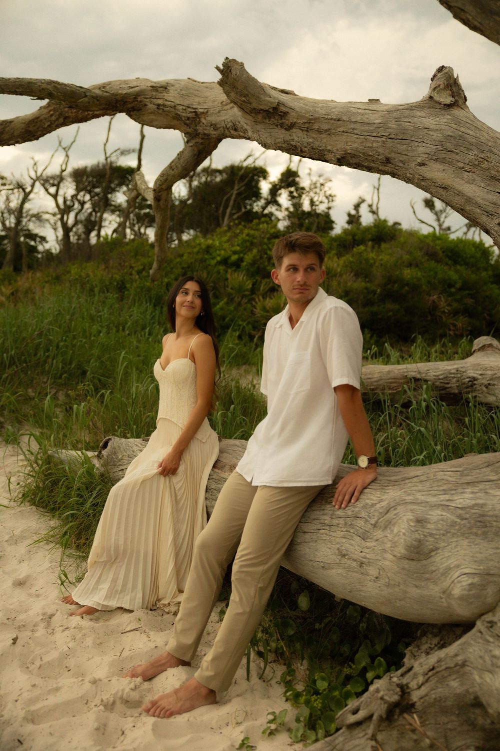 A couple posing by a large driftwood log on a sandy beach, surrounded by lush greenery.